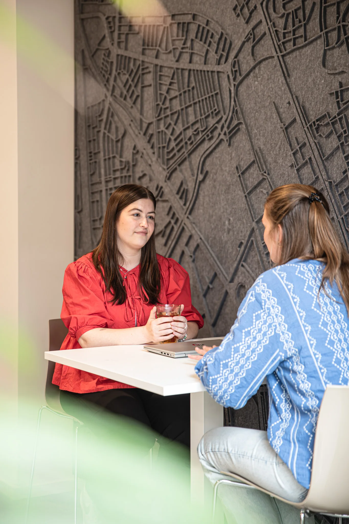 Twee vrouwen zitten tegenover elkaar aan een klein tafeltje en zijn in gesprek. Aan de muur achter hen hangt een reliëfkaart.