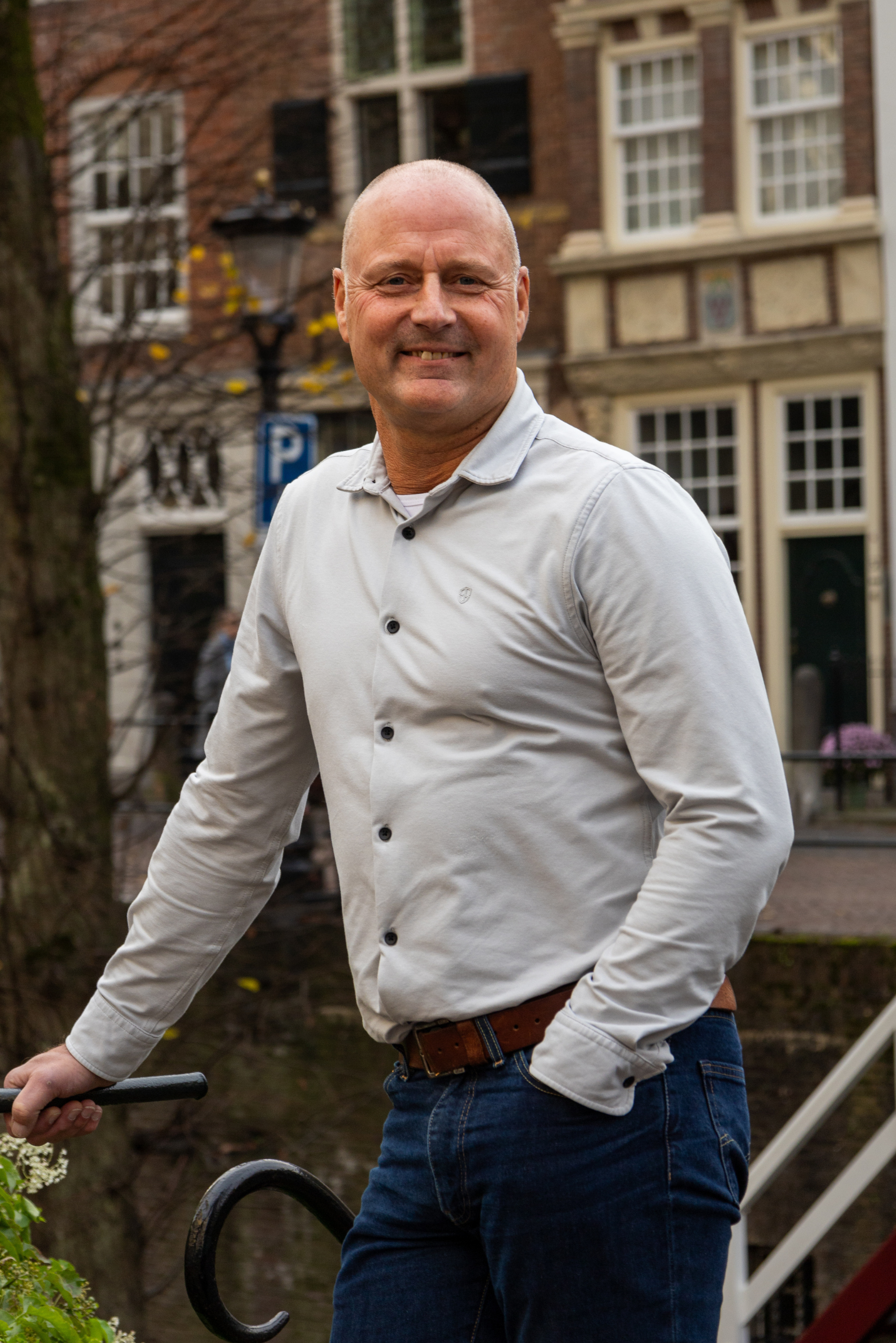 Een man in een wit overhemd staat buiten bij een hek, met gebouwen op de achtergrond.