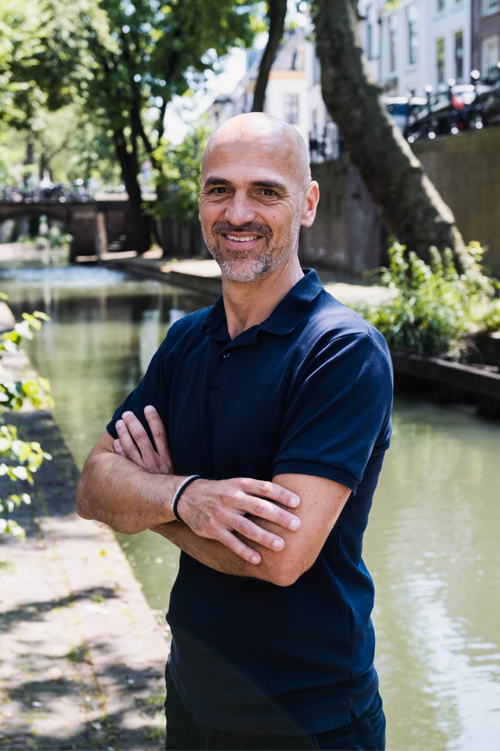 Een kale man die naast een rivier staat met zijn armen over elkaar.