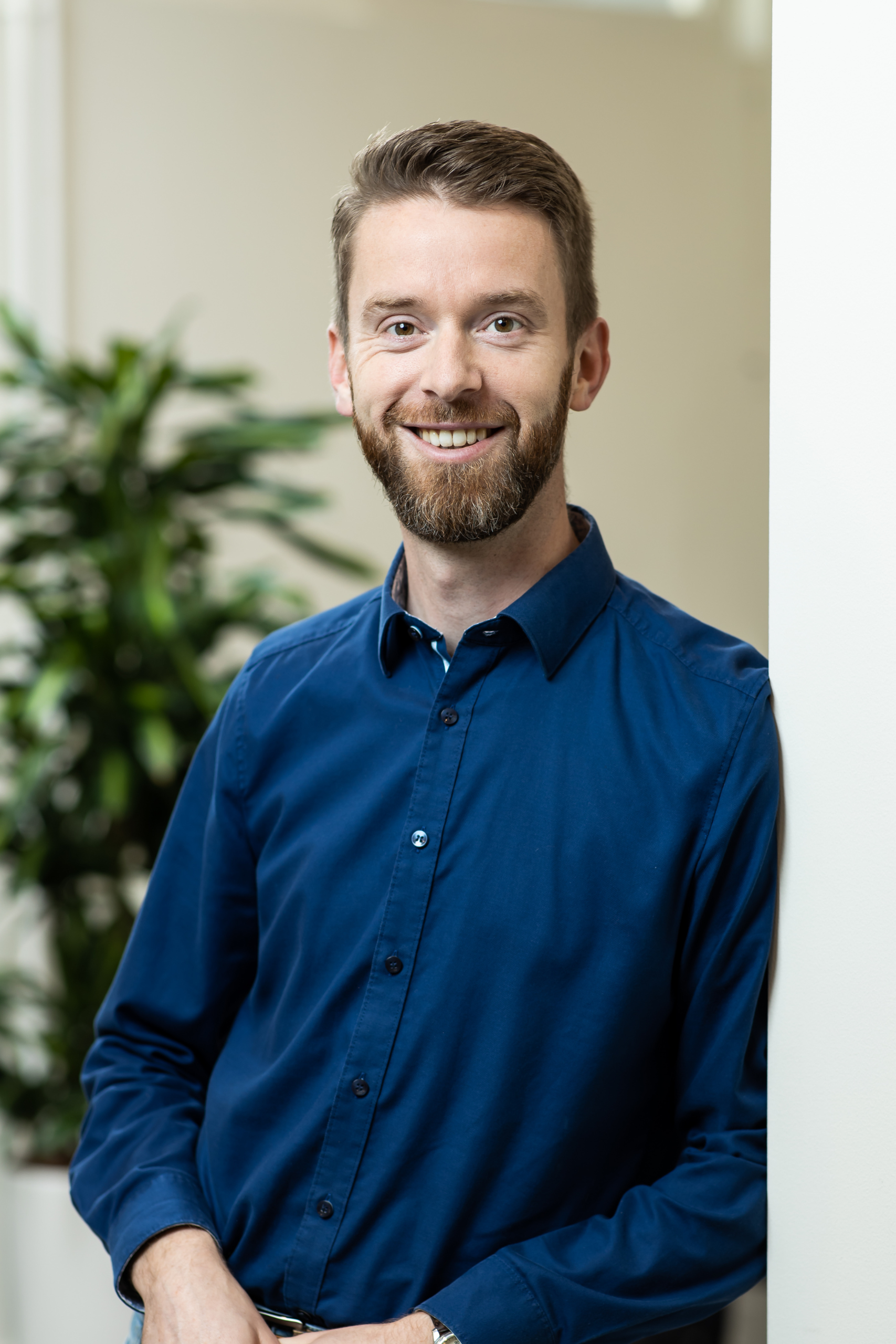 Een bebaarde man in een blauw shirt staat naast een plant.