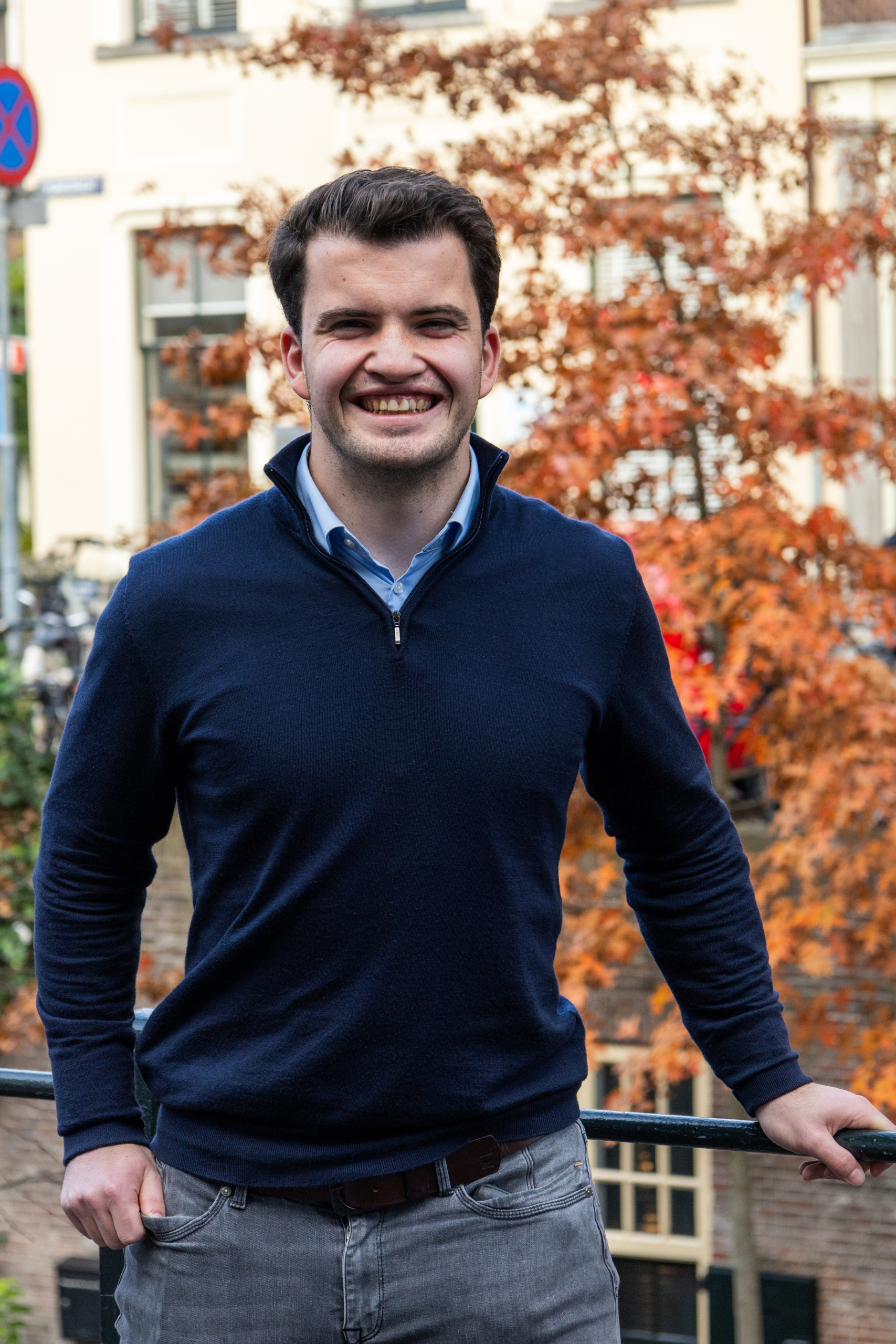 Een persoon in een donkere trui staat buiten, lachend, met herfstbomen op de achtergrond.