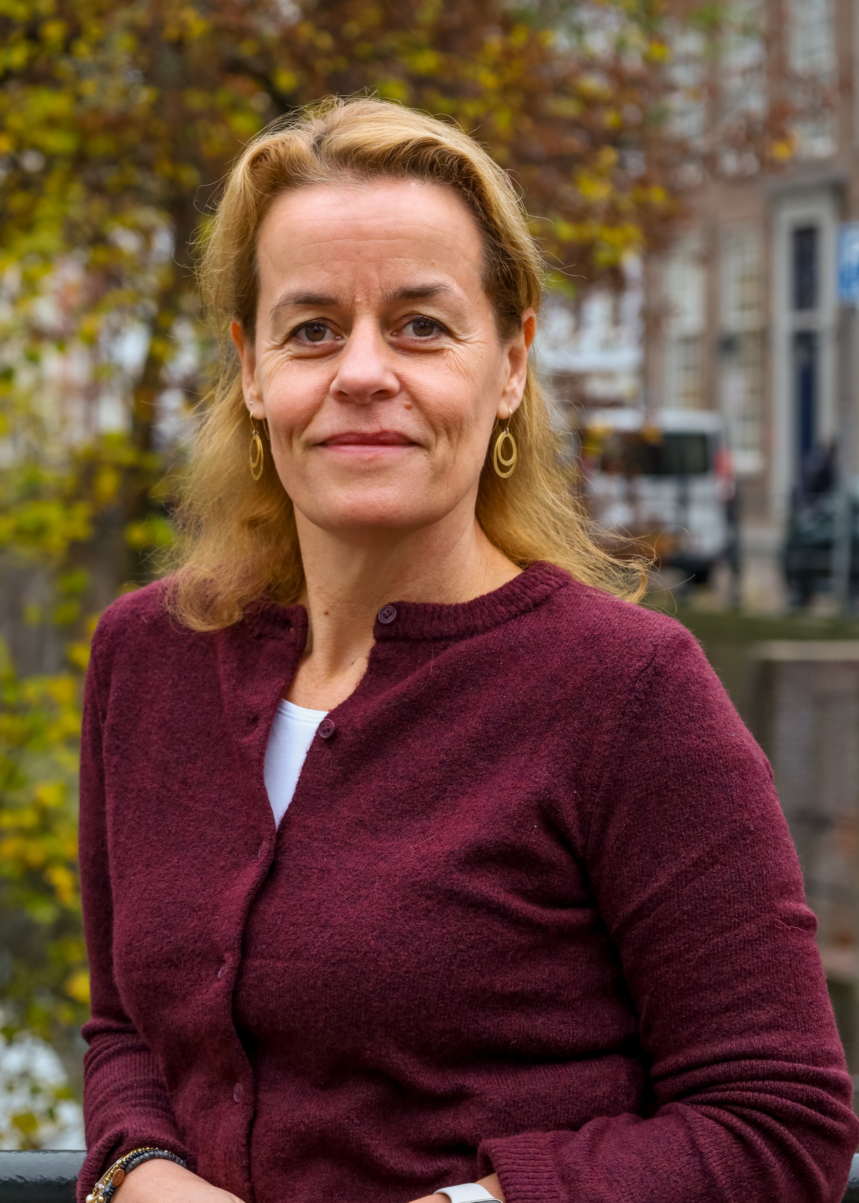 Een vrouw in een kastanjebruine trui staat buiten met wazige gebouwen en herfstbladeren op de achtergrond.