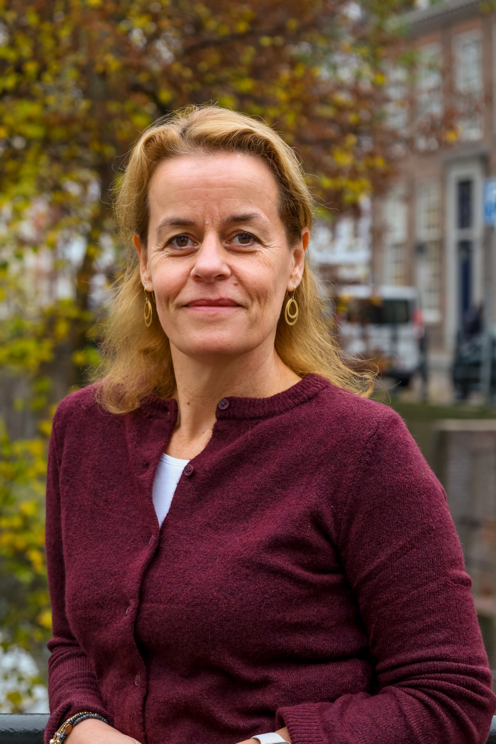 Een vrouw in een kastanjebruine trui staat buiten met wazige gebouwen en herfstbladeren op de achtergrond.