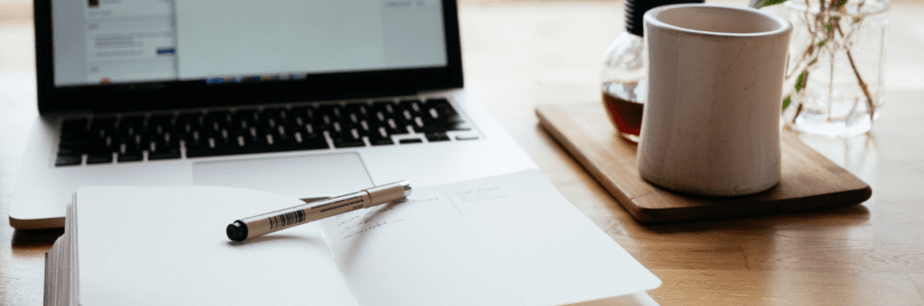 Een notitieboekje met een pen en een laptop op een houten tafel.