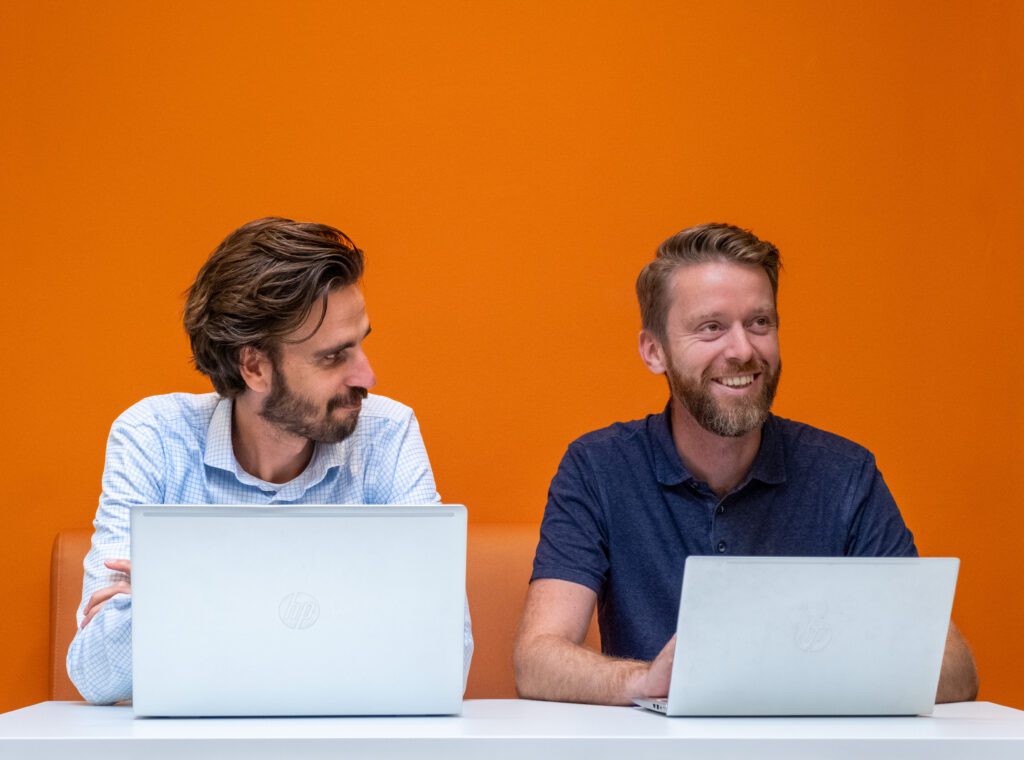 Twee mannen zitten aan een bureau met laptops voor een oranje muur.