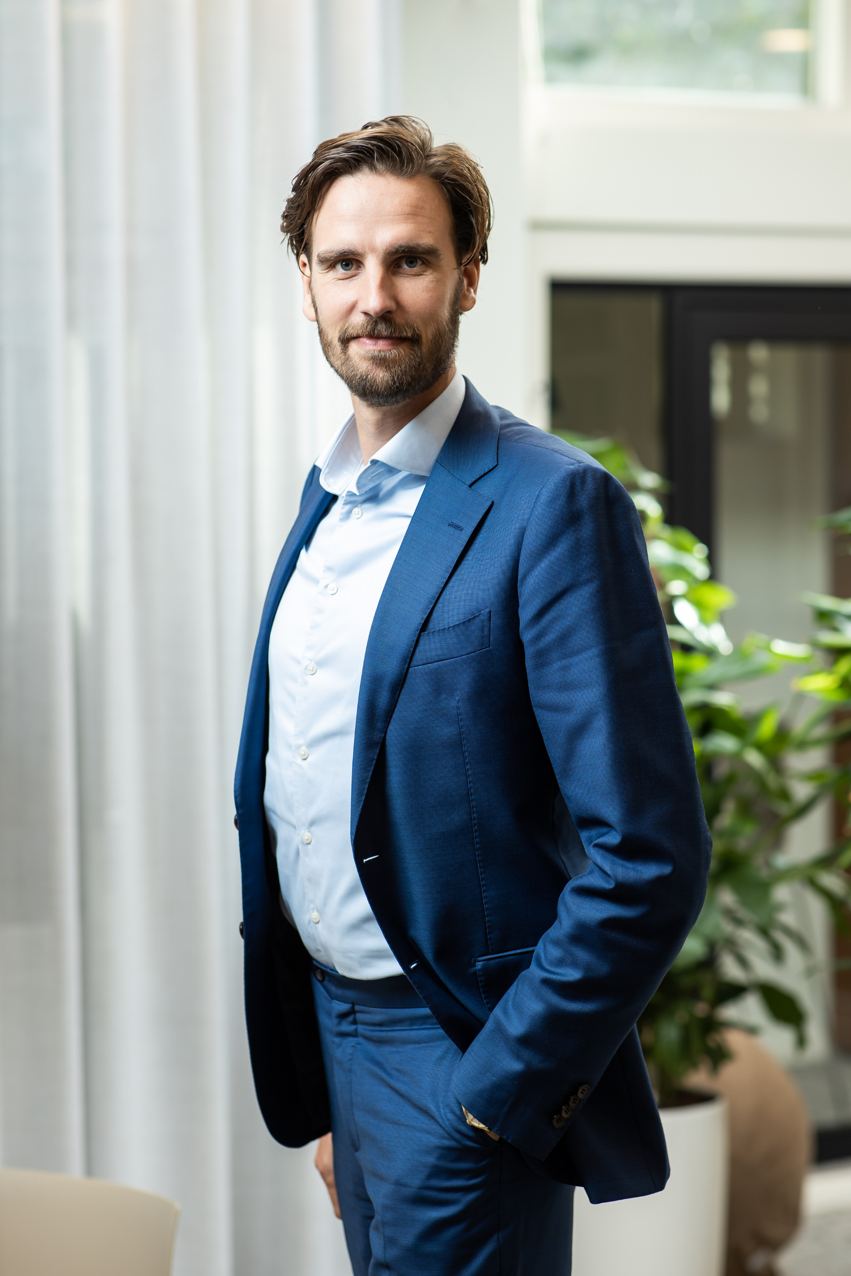 Een man in een blauw pak staat voor een raam.