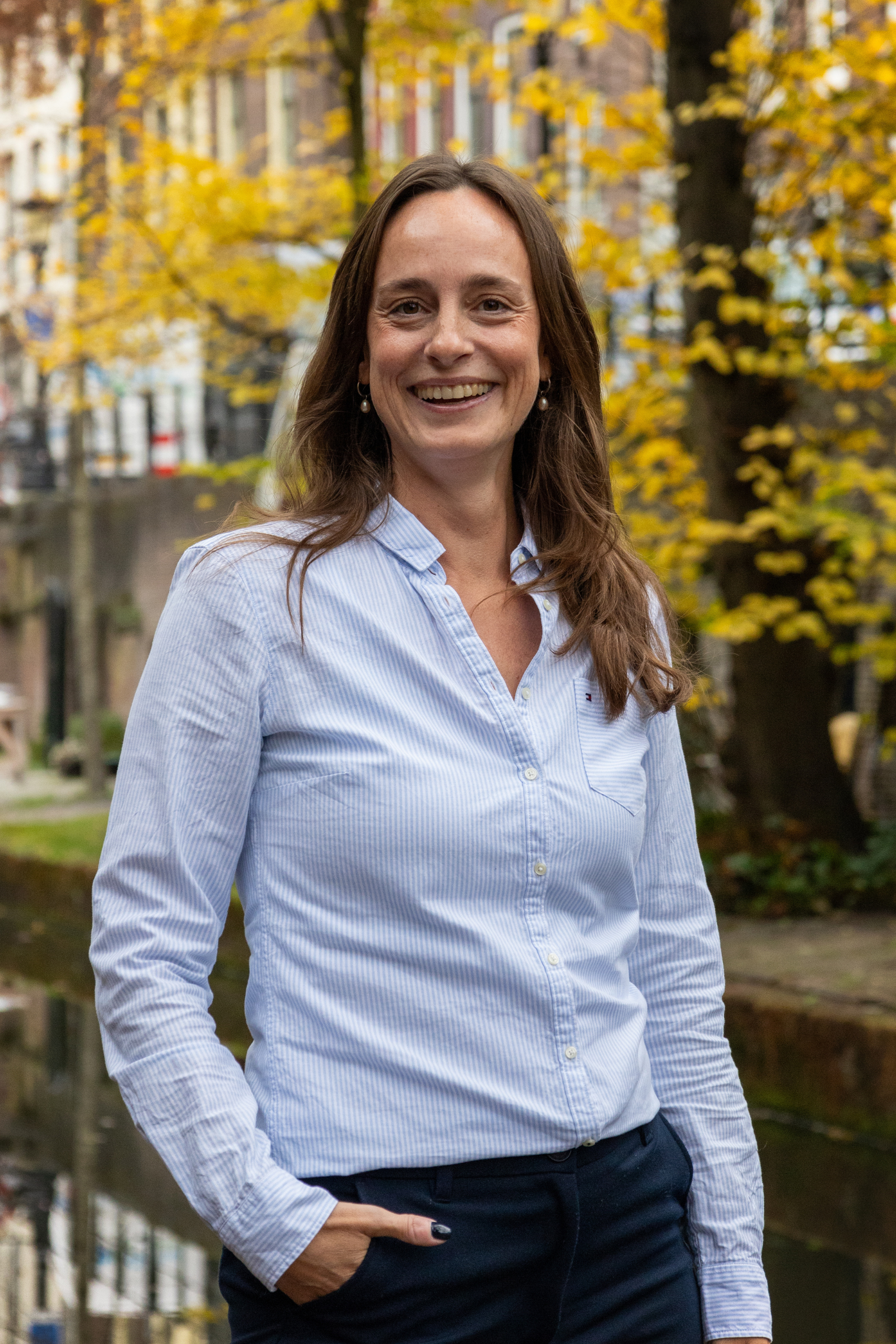 Een persoon in een lichtblauw shirt staat buiten bij een kanaal, met bomen en gebouwen op de achtergrond, en lacht naar de camera.