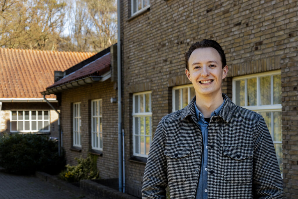 Een persoon staat voor een bakstenen gebouw en lacht zelfverzekerd naar de camera. Als onderdeel van het interim accountmanagementteam van GGz Centraal gaan ze elke uitdaging aan. Bomen en struiken geven de achtergrond een serene sfeer.