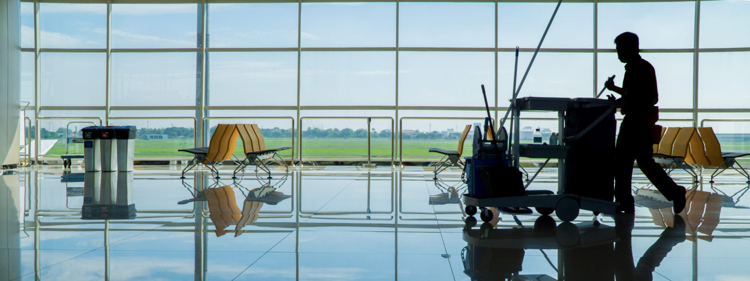 Een silhouet van een persoon die een schoonmaakkar voortduwt over een glimmende vloer in een luchthaventerminal met stoelen en grote ramen op de achtergrond.