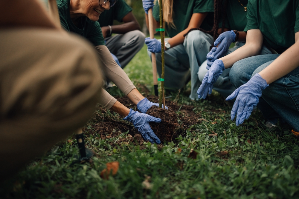 Mensen met blauwe handschoenen aan planten samen buiten een boompje in de grond. Ze doen mee aan een tuin- of boomplantactiviteit in groepsverband.
