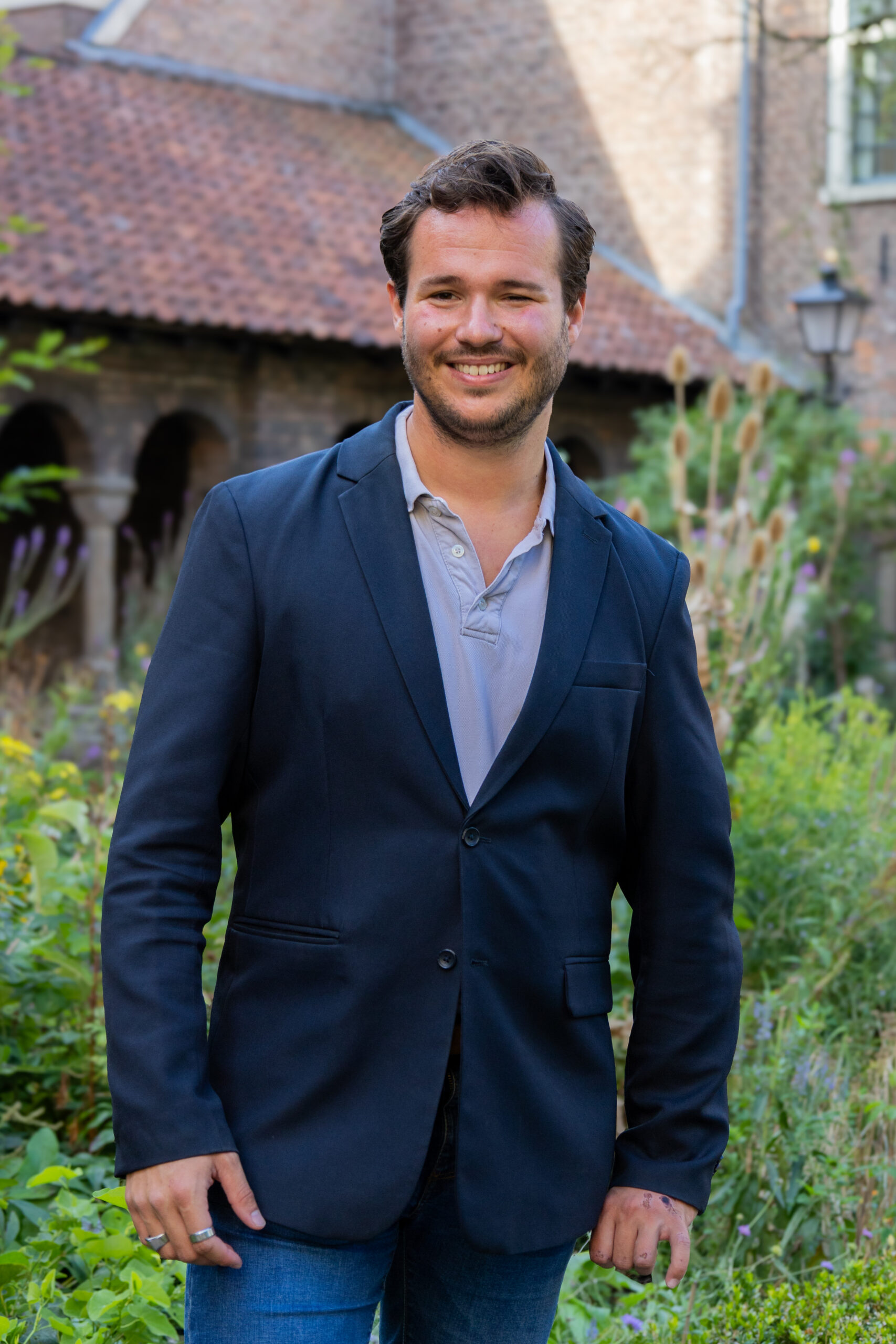 Een man in een donkere blazer en licht overhemd staat te glimlachen in een tuin met groen en een bakstenen gebouw op de achtergrond.