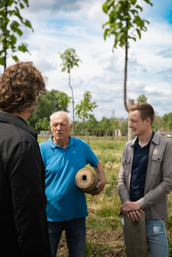 Drie mannen staan buiten in een veld te praten. Eén man in een blauw poloshirt houdt een holle boomstam vast, terwijl de anderen luisteren. Op de achtergrond staan jonge bomen en een bewolkte lucht.
