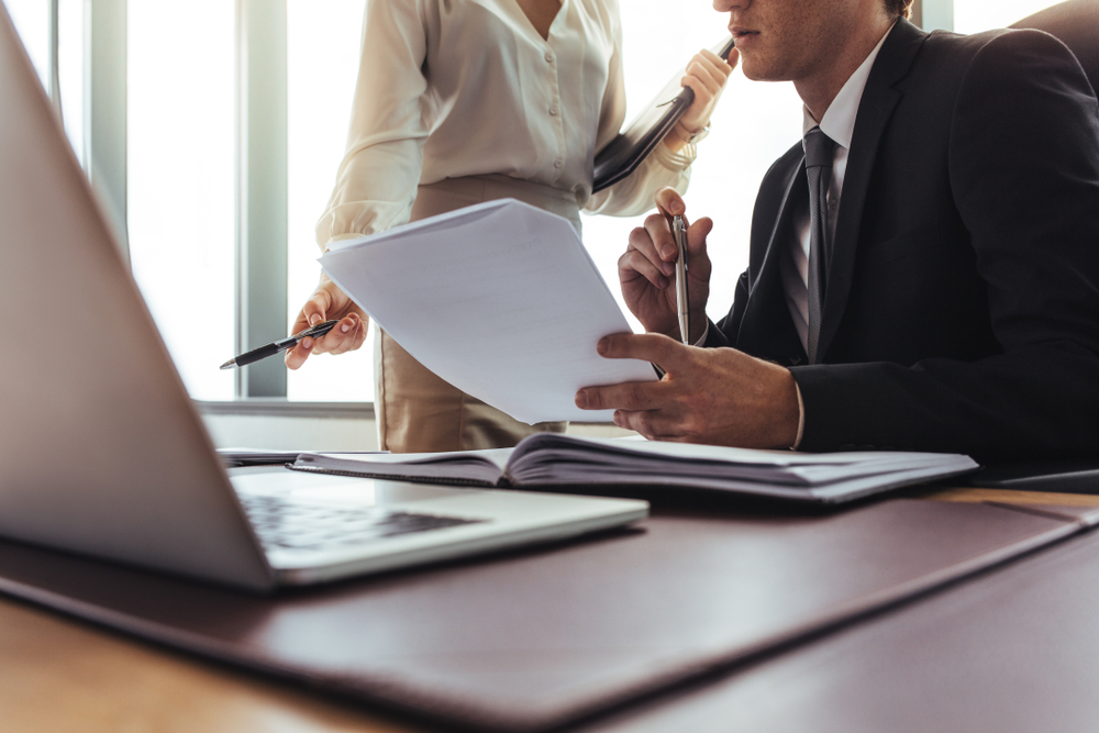 Twee mensen in zakelijke kleding bekijken documenten aan een bureau met een laptop en bestanden in een lichte kantooromgeving.
