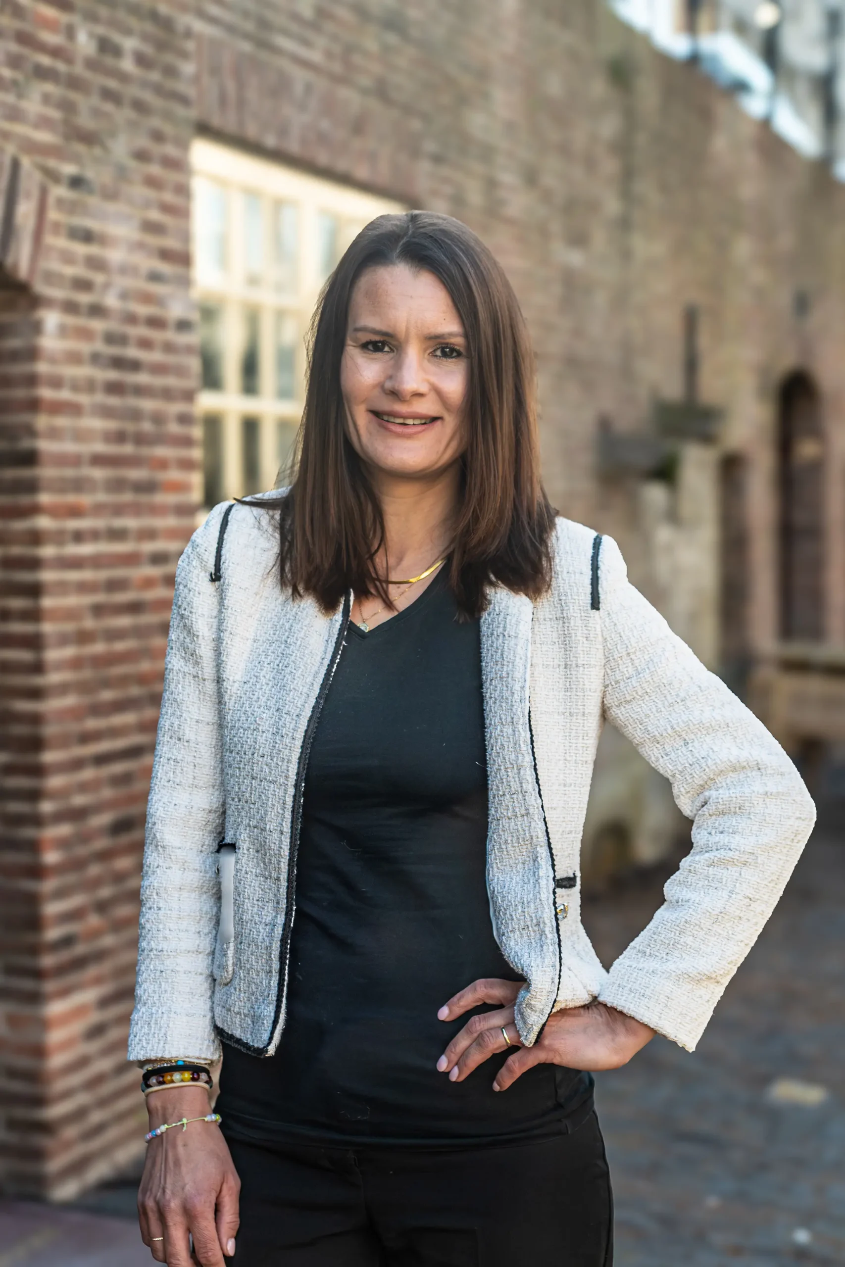 Een vrouw met steil bruin haar staat buiten voor een bakstenen gebouw, ze draagt een wit structuurjasje over een zwart topje en een zwarte broek, met een hand op haar heup.