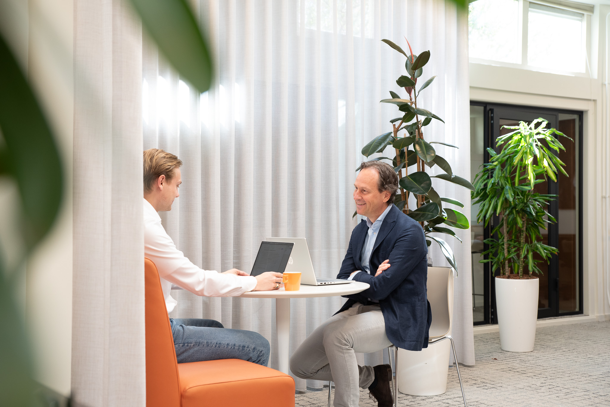 Twee mannen zitten aan een ronde tafel in een lichte kantoorruimte; ze praten en werken op hun laptops. Op de achtergrond staan potplanten en hangen witte gordijnen, waardoor VFM in beeld komt terwijl ze hun nieuwste project bespreken.
